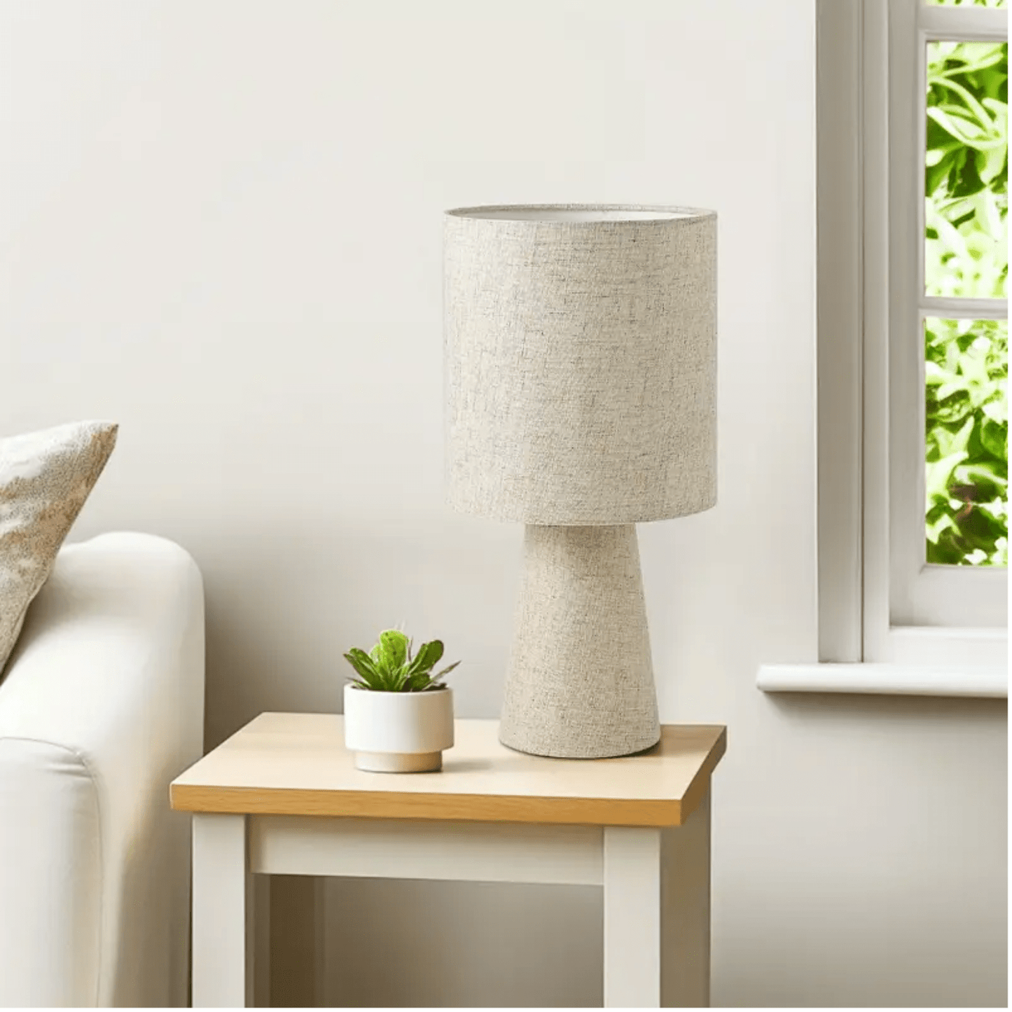 Linnen tafellamp met beige cilindervormige kap op een houten bijzettafel naast een raam met daglicht.