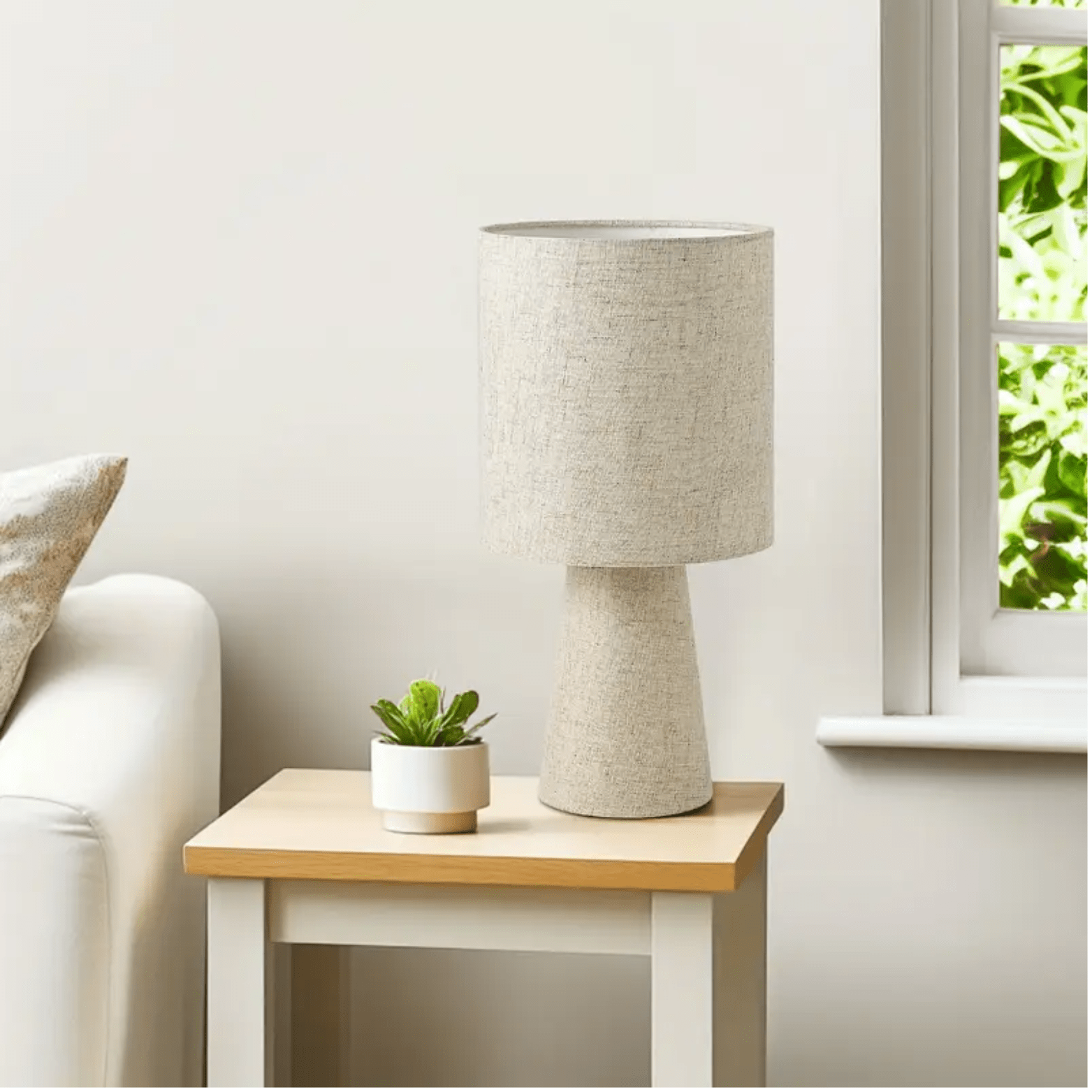 Linnen tafellamp met beige cilindervormige kap op een houten bijzettafel naast een raam met daglicht.