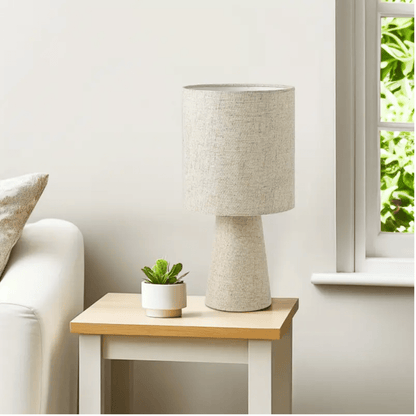 Linnen tafellamp met beige cilindervormige kap op een houten bijzettafel naast een raam met daglicht.