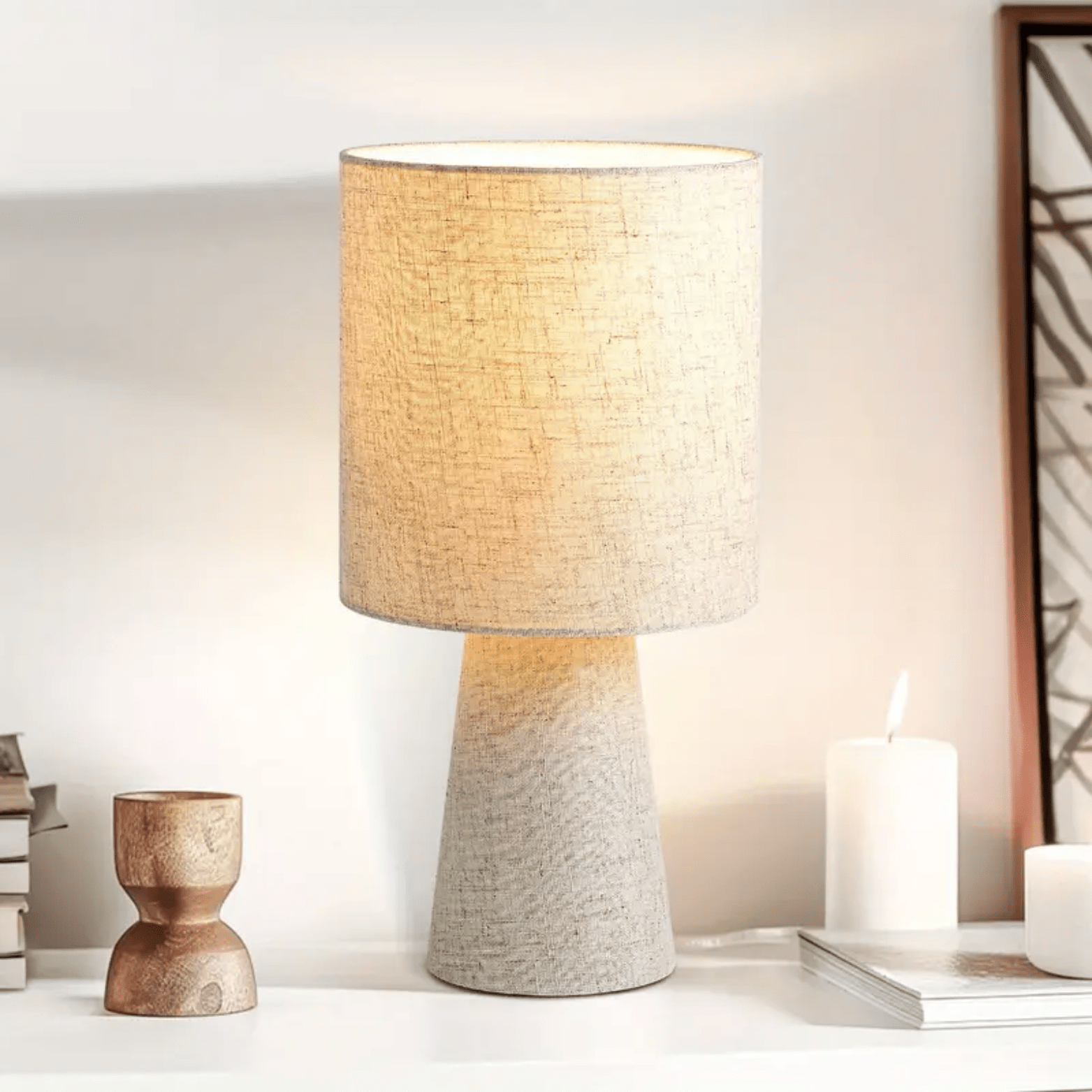 Beige linnen tafellamp met cilindervormige kap op een witte kast naast kaarsen en decoratie.
