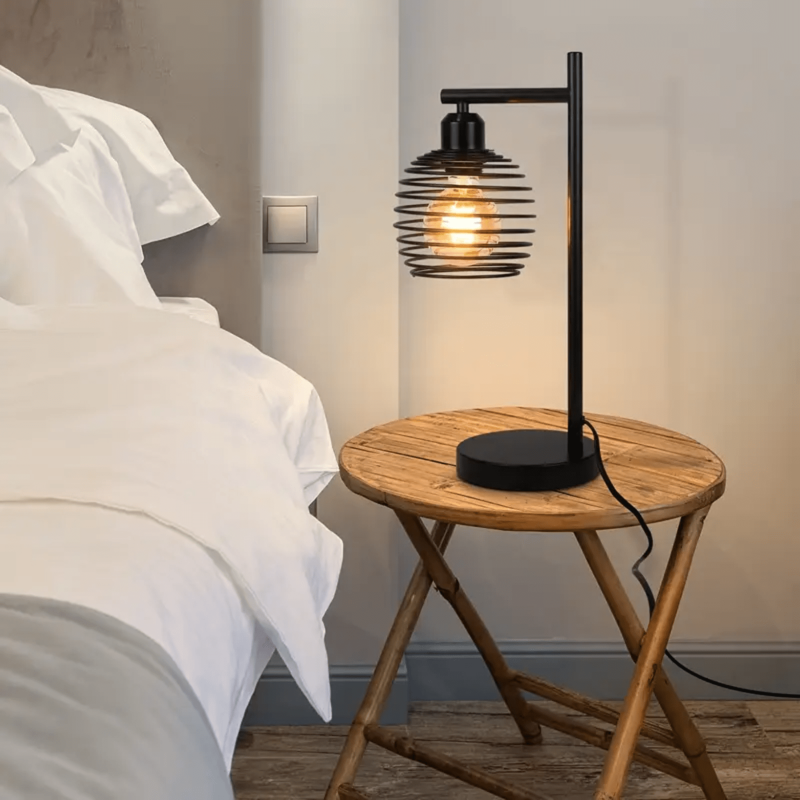 Zwart industrieel nachtlampje met open spiraalkap op rond houten tafeltje naast wit bed.