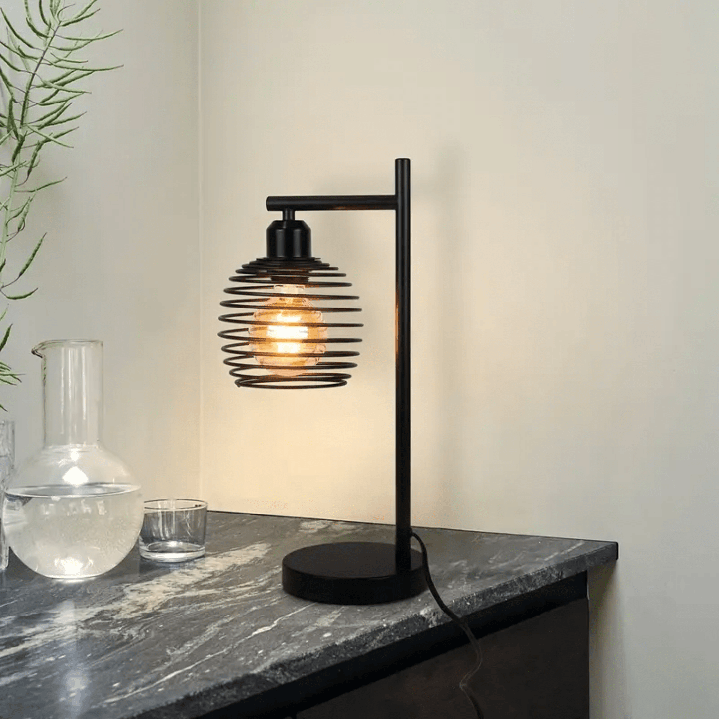 Zwarte industriële tafellamp met open kap en gloeilamp op donker marmeren tafelblad tegen lichte muur.