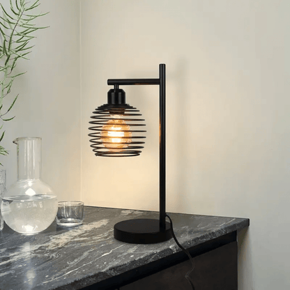 Zwarte industriële tafellamp met open kap en gloeilamp op donker marmeren tafelblad tegen lichte muur.