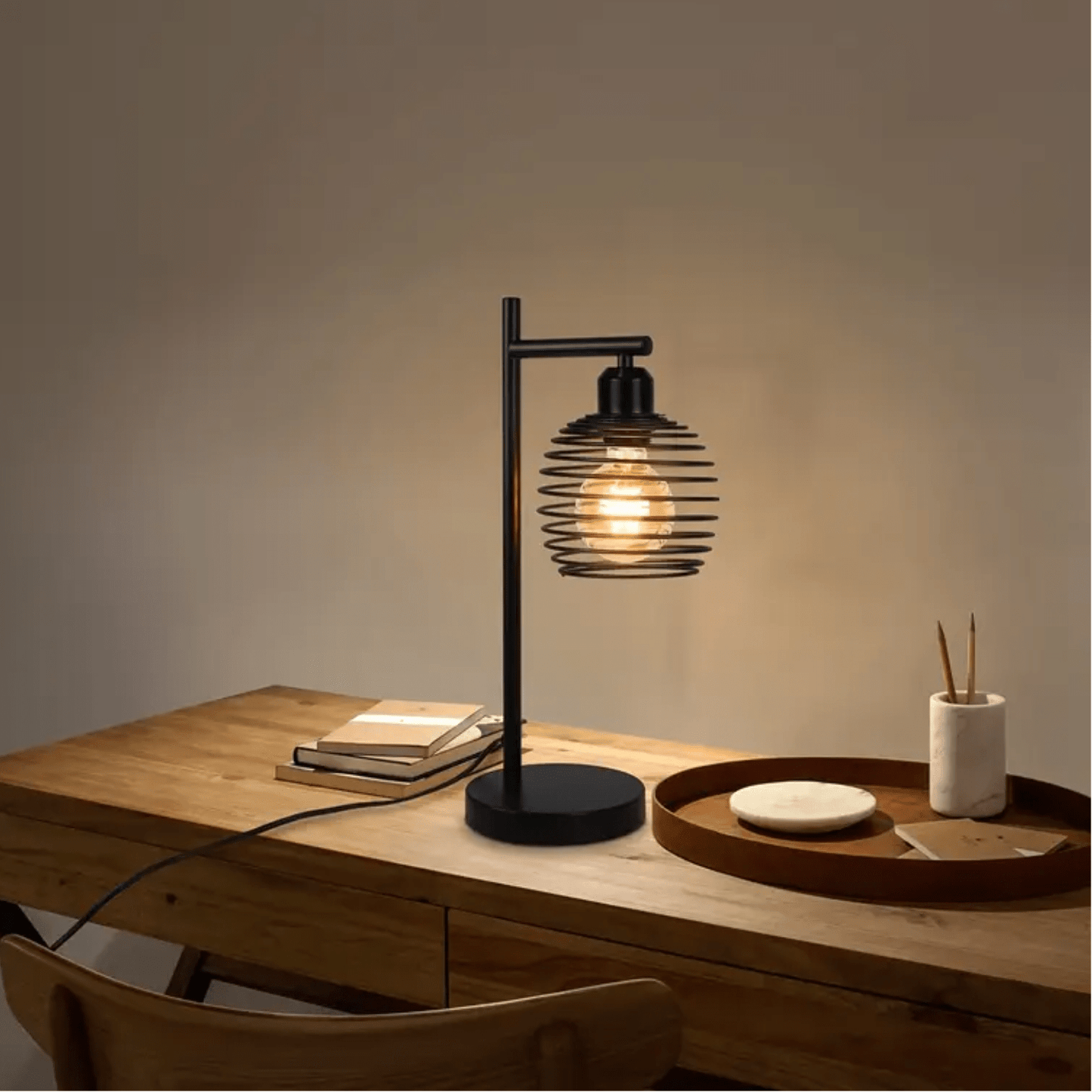 Zwarte tafellamp met spiraalkap en warme gloeilamp op houten bureau met boeken en accessoires.